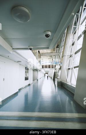 Corridoio moderno lungo del ponte di imbarco dell'aeroporto con pavimento in resina blu, pareti bianche, telecamere a soffitto, grandi finestre sul lato destro e un cartello direzionale bilingue che indica “Centro di trasferimento” in cinese e inglese, situato all'Aeroporto Internazionale di Shanghai Pudong, Cina Foto Stock