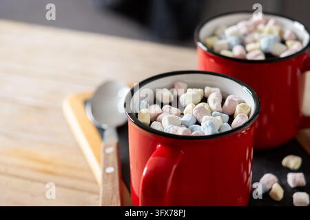 Prima piano di due tazze di ceramica rosse piene di cioccolata calda e mini marshmallow colorati su un tavolo di legno. Foto Stock