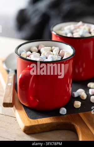Prima piano di due tazze di ceramica rosse piene di cioccolata calda e mini marshmallow colorati su un tavolo di legno. Foto Stock