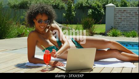 Vista laterale di una giovane donna nera che beve un cocktail mentre utilizza un laptop vicino alla piscina. Si sta rilassando e bevendo un cocktail 4k Foto Stock