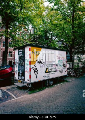 Rimorchio di manutenzione coperto di graffiti parcheggiato sotto gli alberi accanto a biciclette in una strada residenziale ad Amsterdam, Paesi Bassi Foto Stock