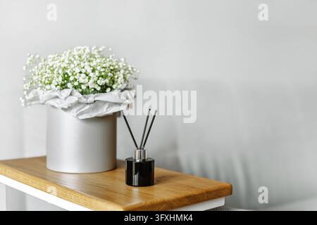 Bottiglia diffusore di aromi in vetro nero e delicate fiori di gypsophila bianchi in una scatola per cappelli in argento posta su un tavolo di legno all'interno di una stanza accogliente. Minima Foto Stock
