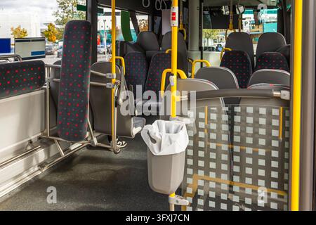 L'interno di un autobus moderno presenta file di sedili grigi e neri con motivi rossi. Un cestino della spazzatura bianco è attaccato a un palo verticale giallo nea Foto Stock