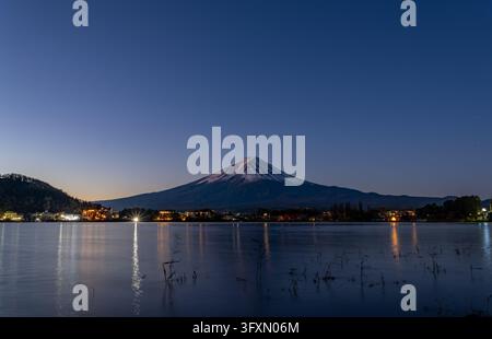 Il monte Fuji coperto di neve si erge sopra una città con edifici illuminati, riflessi nelle acque calme di un lago sotto un cielo notturno stellato. Foto Stock