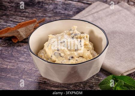 Primo piano di gelato stracciatella servito in una ciotola di ceramica beige accanto a un tovagliolo di lino piegato, corteccia di cannella e rametti di menta su legno stagionato. Ghiaccio italiano Foto Stock