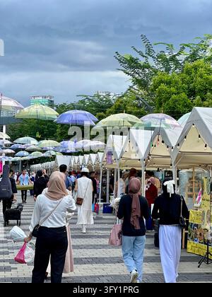 Persone che camminano lungo un mercato di strada all'aperto con ombrelli sospesi trasparenti. I pedoni passeggiano attraverso una strada di mercato all'aperto fiancheggiata da bancarelle di tè bianche Foto Stock