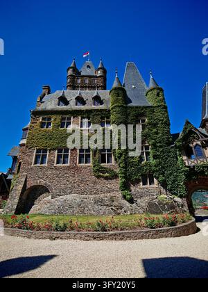 Facciata maestosa del castello medievale di Reichsburg Cochem con mura di pietra ricoperte di edera, guglie, bandiera e giardino di rose in Germania. Foto Stock