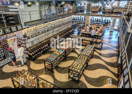I visitatori esplorano un negozio di ceramiche e piastrelle a Porto, Portogallo. Il pavimento mostra un motivo ondulato di aree scure e chiare. Magneti e bottoni di ceramica sono disposti sui tavoli di vendita, e immagini di piastrelle sono appese alle pareti. Gli azulejos sono piastrelle di ceramica dipinte e smaltate. La parola deriva dall'arabo "al-zillīj" (pietra levigata). Foto Stock