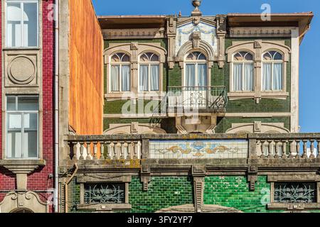 Una facciata di casa con piastrelle verdi, finestre ad arco rotondo e un balcone in ferro battuto si trova a Porto, Portogallo. Un fregio di azulejos con un vaso e ornamenti decora il parapetto. A sinistra si affianca una facciata in mattoni rossi. La produzione industriale di piastrelle per facciate iniziò nella città intorno al 1840. La moda di coprire le case con azulejos venne dal Brasile. Foto Stock