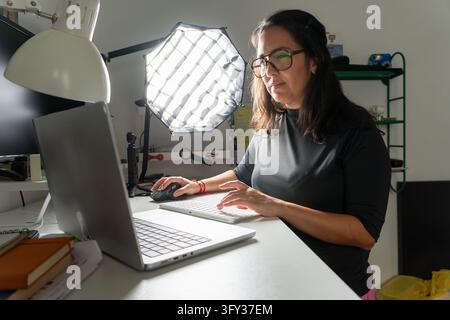 Donna concentrata che lavora sul suo laptop a una scrivania con una tastiera e un mouse esterni, illuminata da luci da studio Foto Stock