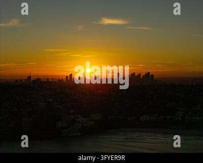 Vista aerea dello skyline di Sydney silouettato contro un vivace tramonto dorato con il sole che tramonta dietro i grattacieli della città e le acque del porto a Sydney Foto Stock