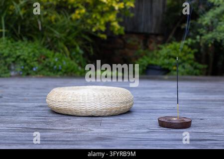 Cuscino rotondo intrecciato color crema appoggiato accanto a un bastoncino d’incenso acceso su un terrazzo di legno, il fumo si alza. Consapevolezza, meditazione, relax, zen, aromaterapia, benessere Foto Stock