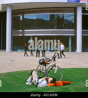 Un uomo ciclista prende una pausa rilassante sull'erba con gli imprenditori a piedi passato edificio per uffici in abiti Moorgate City di Londra UK KATHY DEWITT Foto Stock