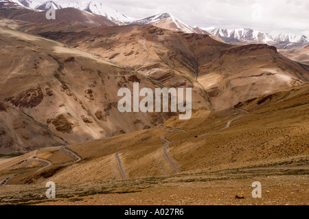 Strada tortuosa attraverso formazioni rocciose rossastre al passo di Taglangla sull'autostrada Manali-Leh a Ladakh, India. Montagne himalayane innevate alle spalle. Foto Stock