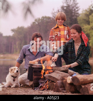 Marito e moglie e figlio per la cottura su un falò con il loro cane accanto a un lago in background Foto Stock