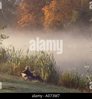 Uomo seduto da solo accanto canne di prima mattina canna da pesca e nebbia coperta lago autunno alberi nella campagna Essex Weald Country Park Brentwood Inghilterra UK Foto Stock