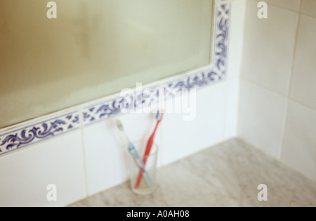 Una coppia defocussed di spazzolini da denti in un bicchiere su un ripiano di marmo o banco sotto una appannata specchio Foto Stock