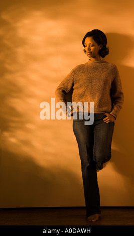 A 30 s African American donna appoggia contro una parete profondo nel pensiero e scalzi in abbigliamento casual Foto Stock
