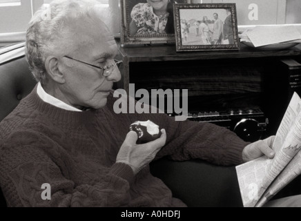 Anziano uomo in pensione a casa leggere la pagina finanziaria del giornale e mangiare una mela durante il blocco del coronavirus. La vita a casa da sola Foto Stock