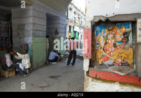 India Dehli Connaught place barbiere al lavoro e la gente che parlava sotto le arcate con poster di hindi scena religiosa in frgd Foto Stock