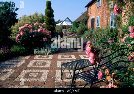 Le rose rosa intorno a piazza Ghiaia e pavimentazione in mattoni nella parte anteriore del cottage. Mobili di metallo. Ritagliato conifera albero in background Foto Stock