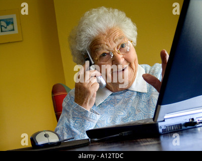 Anziana anziana e vivace donna sul computer è affascinato e stupito dallo schermo del suo computer portatile e parlando sul suo cellulare telefono Foto Stock