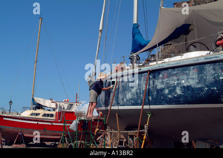 Un uomo che indossa un ampio colmato hat sfrega verso il basso dello scafo di una barca in una barca cantiere di riparazione Foto Stock