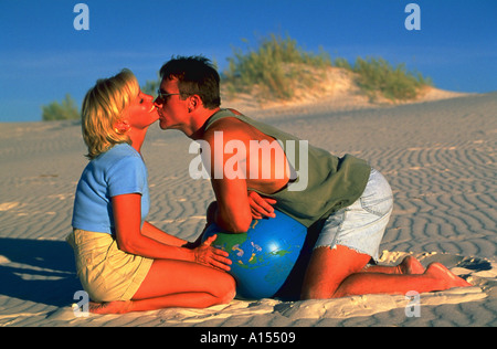 Un uomo e una donna in ginocchio in sabbia entre l'uomo bacia la donna la guancia e si affaccia su una spiaggia di palla che sembra un globo Foto Stock