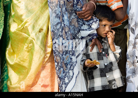 Giovane ragazzo indiano in piedi in una coda tra le donne indossano sari di mangiare un biscotto. Andhra Pradesh, India Foto Stock