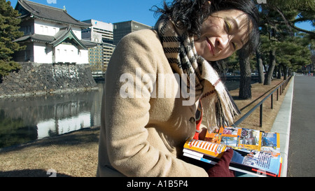 Donna Giapponese in cappotto e sciarpa seduto e guardando la telecamera durante la lettura di una guida a Tokyo giardini del Palazzo Imperiale del Giappone Foto Stock