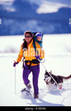 Una giovane madre di racchette da neve con i suoi sei mesi di vecchio figlio sulla sua schiena seguito dal suo cane Husky Foto Stock