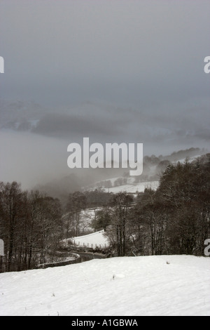 Coperta di neve terreni agricoli con un avvolgimento country road e nebbioso colline innevate in inverno nel Lake District inglese Foto Stock
