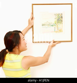 Vista posteriore di una giovane donna il montaggio di una cornice immagine sulla parete e sorridente Foto Stock