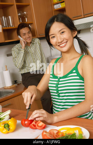 Ritratto di una giovane donna di tagliare le verdure in cucina con un giovane uomo che parla di un telefono cellulare in background Foto Stock