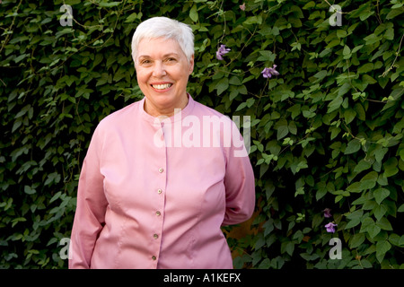 Un allegro e sorridente senior citizen donna sorge nella parte anteriore dei cespugli verdi guardando molto felice. Foto Stock