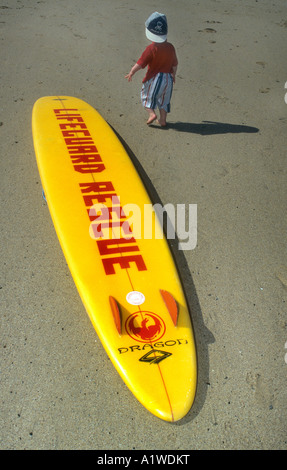 Bagnino di salvataggio con la tavola da surf giacente su di una spiaggia di sabbia in Newquay Cornwall,con un ragazzino passato a piedi. Foto Stock