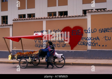 Femmina commerciante di strada che porta il bambino sulla schiena spingendo coperto triciclo di stallo di frutta oltre la parete con graffiti il messaggio di amore Foto Stock