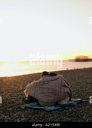 Giovane avvolto in una coperta seduto sulla spiaggia, guardando il tramonto, vista posteriore Foto Stock