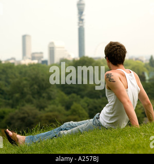 Giovane uomo si siede su Primrose Hill con la skyline di Londra in background in estate Foto Stock