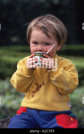 Close-up di adorabili poco ragazzo seduto fuori a mangiare il budino cup e facendo un pasticcio Foto Stock