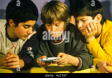 Un giovane ragazzo sta giocando con il suo gameboy mentre i suoi due amici stanno guardando interestedly Foto Stock