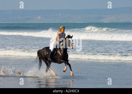 Indonesia Bali Seminyak donna in abito bianco a cavallo lungo la spiaggia Foto Stock