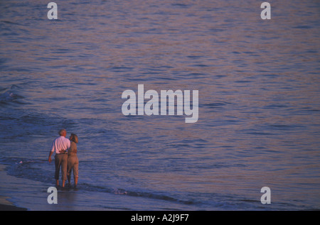 Casualmente vestito coppia matura il sorriso a vicenda per camminare insieme attraverso il dolce profondo della caviglia surf al tramonto Foto Stock