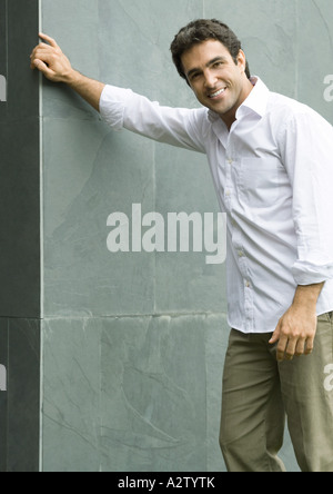 Uomo in piedi con la mano sul muro, sorridente in telecamera, ritratto Foto Stock