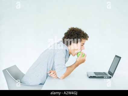 Giovane uomo seduto a tavola, mangiare e apple utilizzando laptop Foto Stock