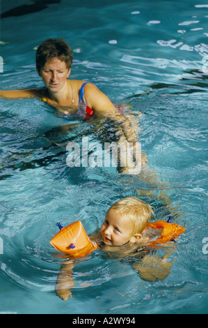 Una giovane madre aiuta il suo bambino a imparare a nuotare in una piscina indossando le bande del braccio Foto Stock