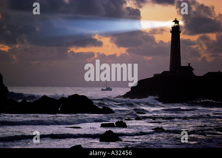 Fasci di luce che si irradia da Pigeon Point lighthouse attraverso il cielo nuvoloso sopra shoreswith rocciose barca da pesca in California a distanza Foto Stock