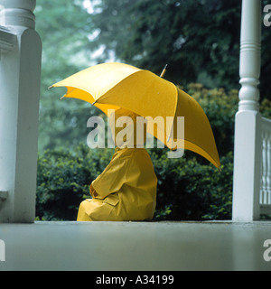 Un bambino che indossa un giallo cappotto di pioggia e stivali da pioggia in piedi sotto la pioggia tenendo un ombrello giallo Foto Stock