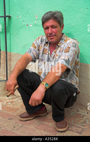 Uomo di fumare un sigaro, accovacciata e poggiante contro una parete, Trinidad, Sancti Spiritus Provincia, Cuba Foto Stock