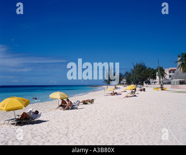 Caraibi Barbados Indie ad ovest della costa sud di Dover turisti in spiaggia al sole su una sedia a sdraio sotto la pantina parasole ombrelli sulla sabbia bianca Foto Stock